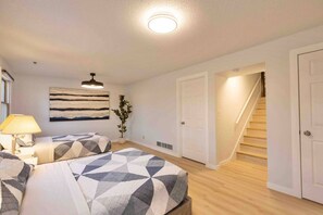 A bedroom with two beds, a nightstand, a lamp, a ceiling light, a wall art piece, and a staircase leading to another room.