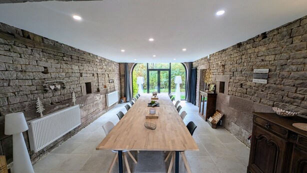 Une salle à manger avec une longue table en bois, des murs en pierre et une vue dégagée grâce à de grandes fenêtres.