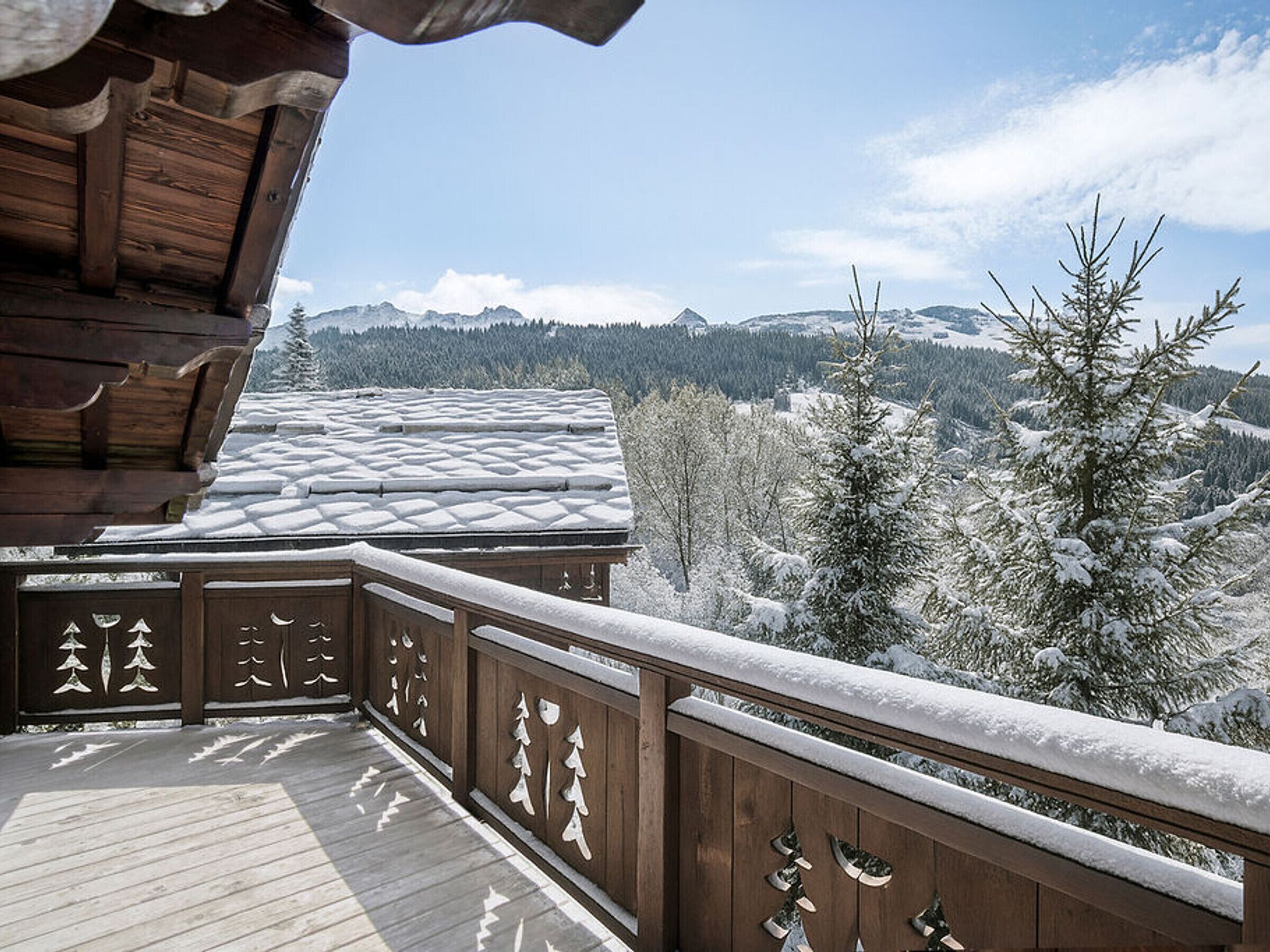A wooden deck with snow-covered railings and a view of snow-covered trees and mountains.