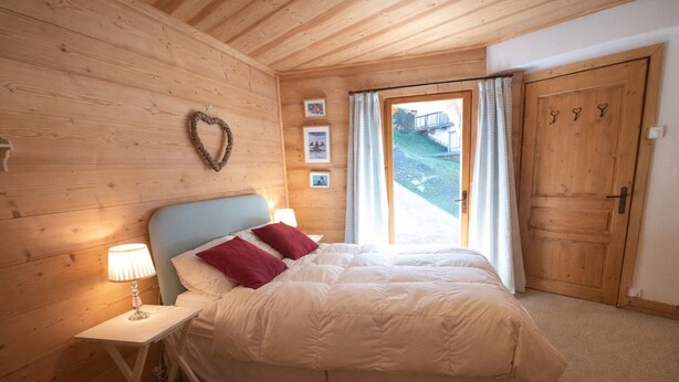 Quarto com teto de madeira, cama com roupa de cama branca e almofadas vermelhas, mesa de cabeceira com abajur, decoração em formato de coração na parede e porta com ganchos em formato de coração.