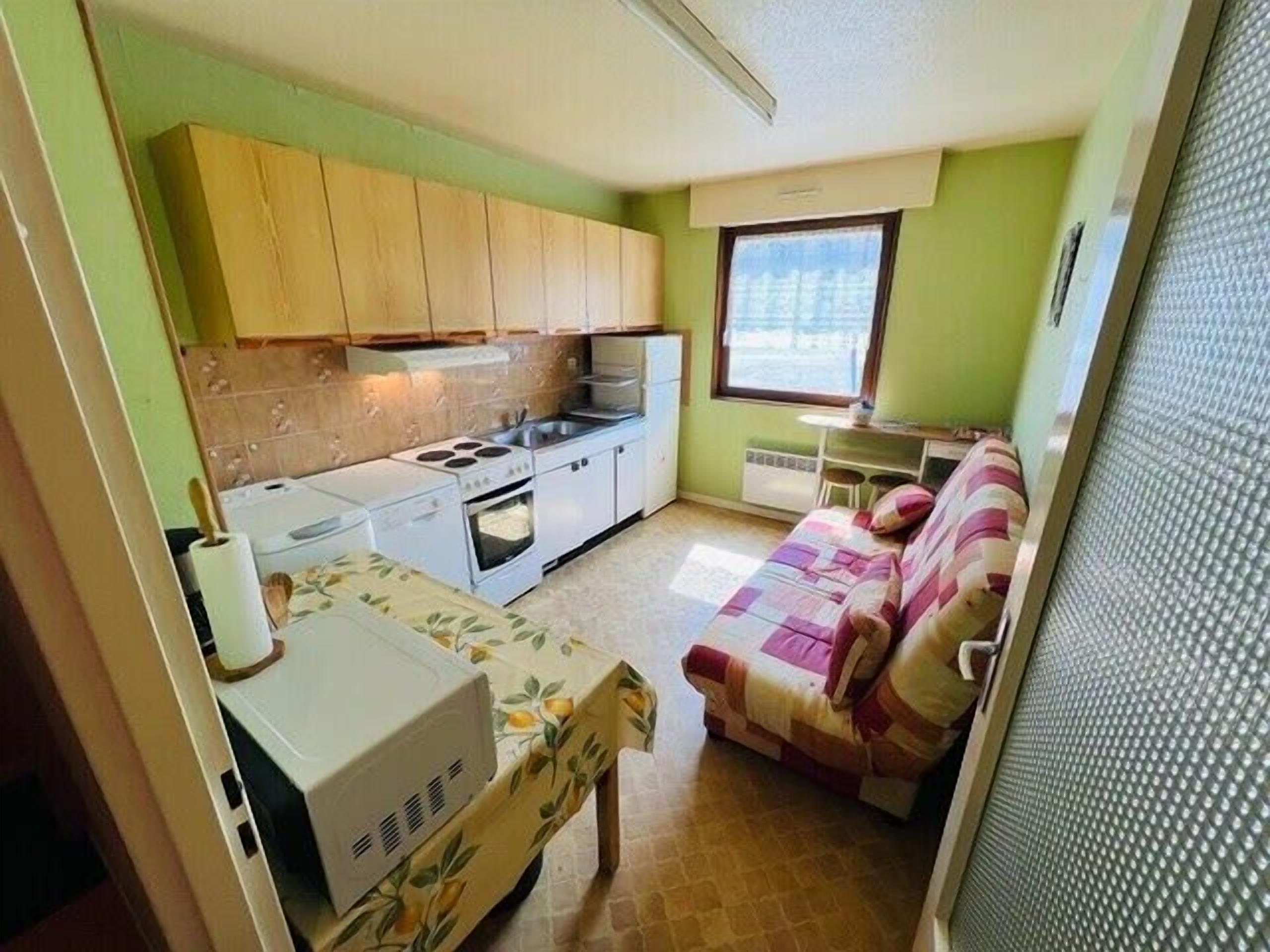 A compact kitchen with a stove, oven, and sink, a small dining table with a floral tablecloth, and a sofa with a patterned cover.