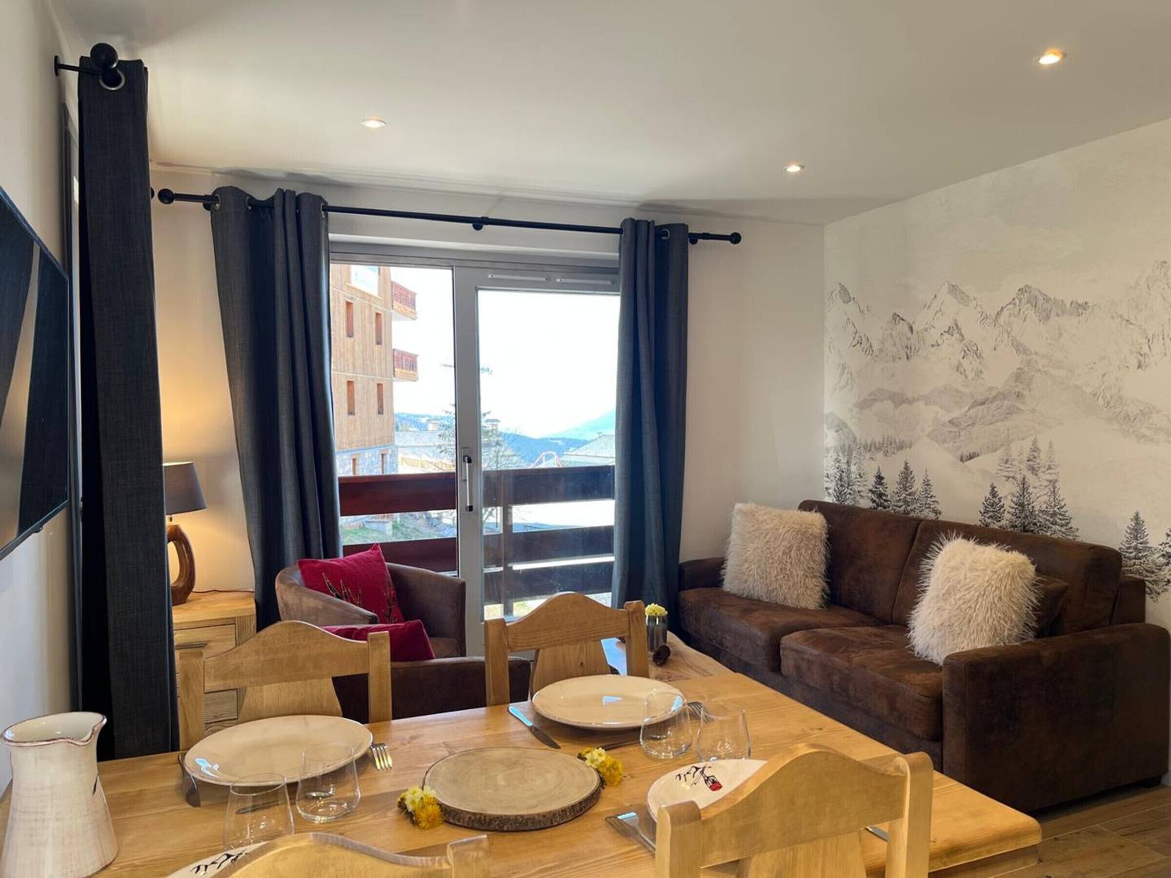 Un salon avec une table à manger, un canapé marron agrémenté de coussins, une fenêtre avec des rideaux et une vue sur la montagne.