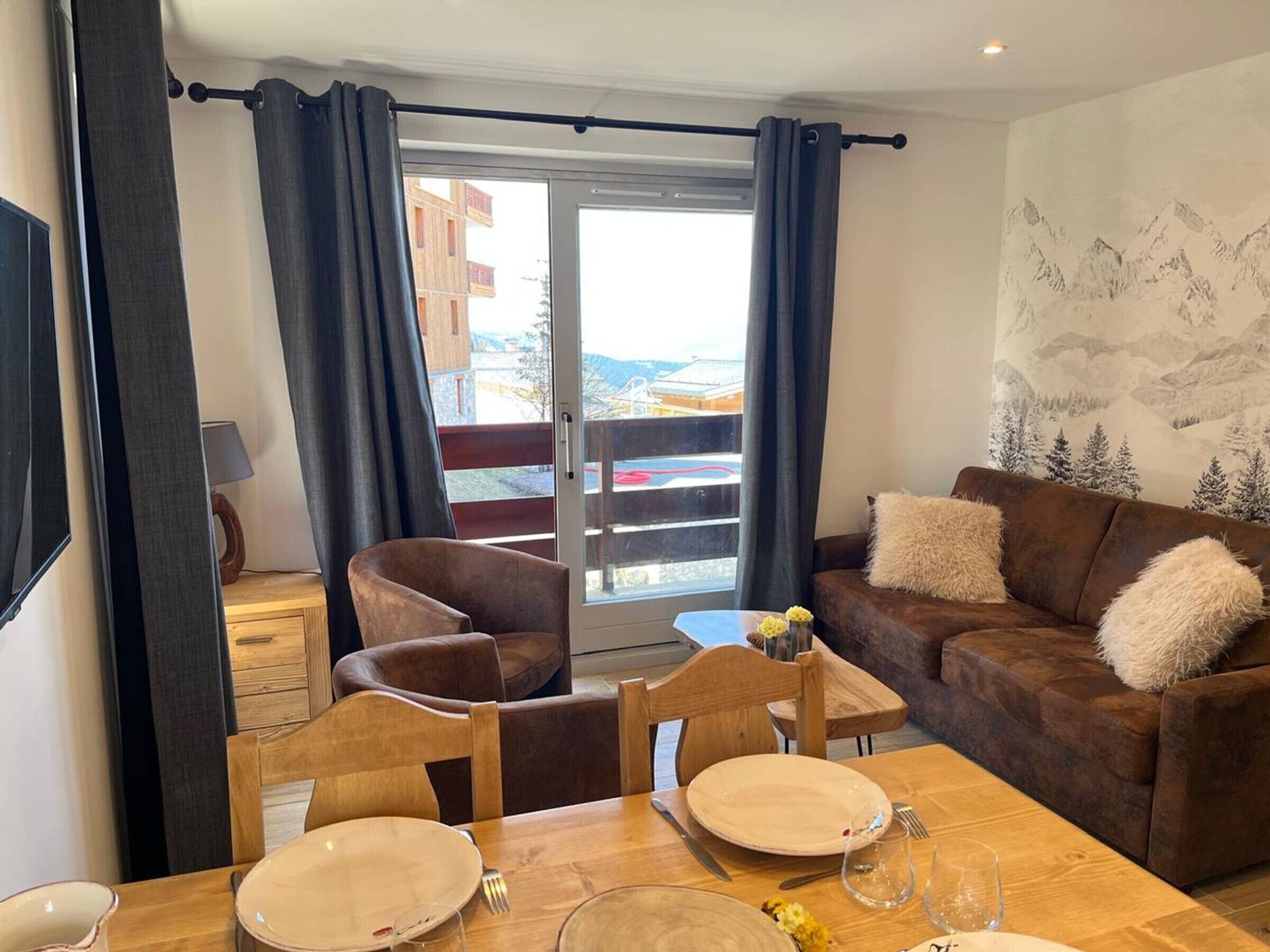 A dining area with a table set for two, a brown sofa with fluffy pillows, and a balcony with a view of buildings and mountains.