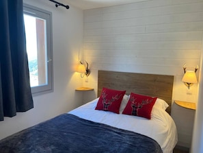 A bedroom with a bed, two wall-mounted lamps, a window with curtains, and a view of the outdoors.