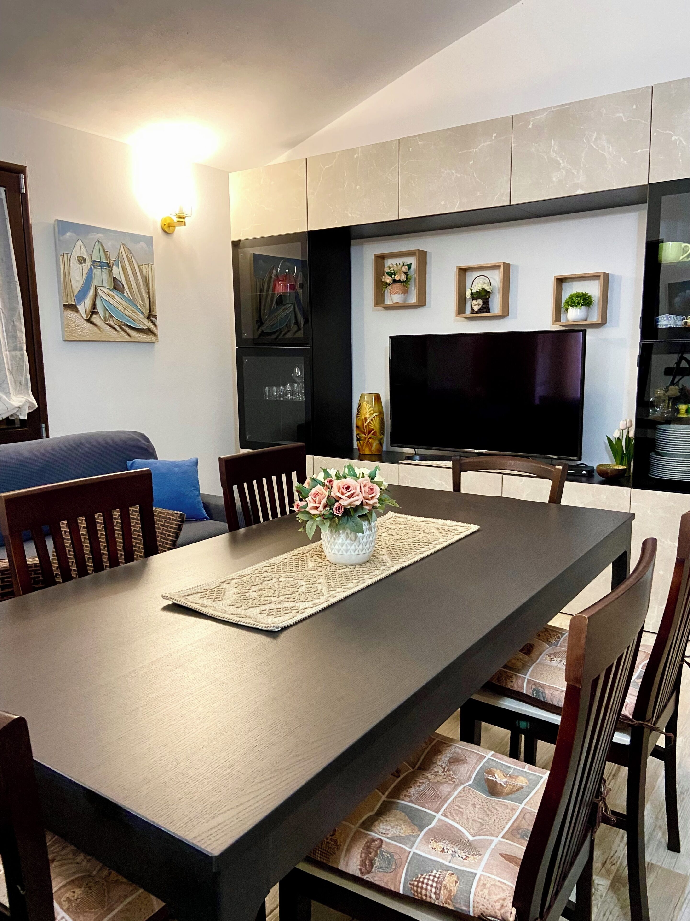 Un comedor con una mesa larga, sillas, un jarrón con flores, un televisor y estanterías con objetos decorativos.