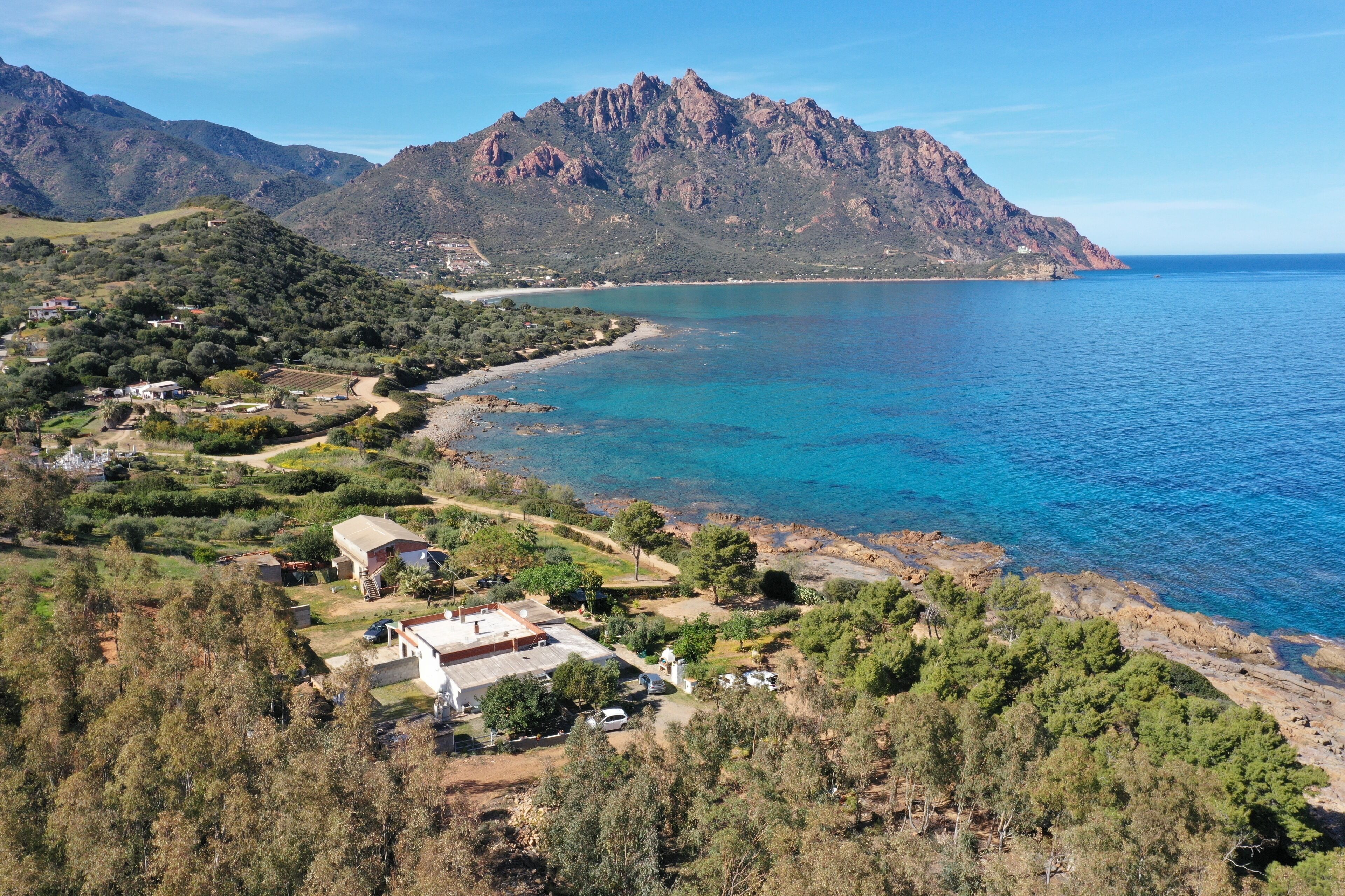 A coastal area with a mountain in the background, a clear blue sea, and a few buildings with visible structures and greenery.