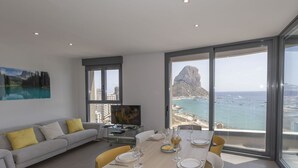 Un salon moderne comprenant un coin repas, de grandes fenêtres offrant une vue sur la mer et les montagnes, un canapé avec des coussins et un téléviseur à écran plat.