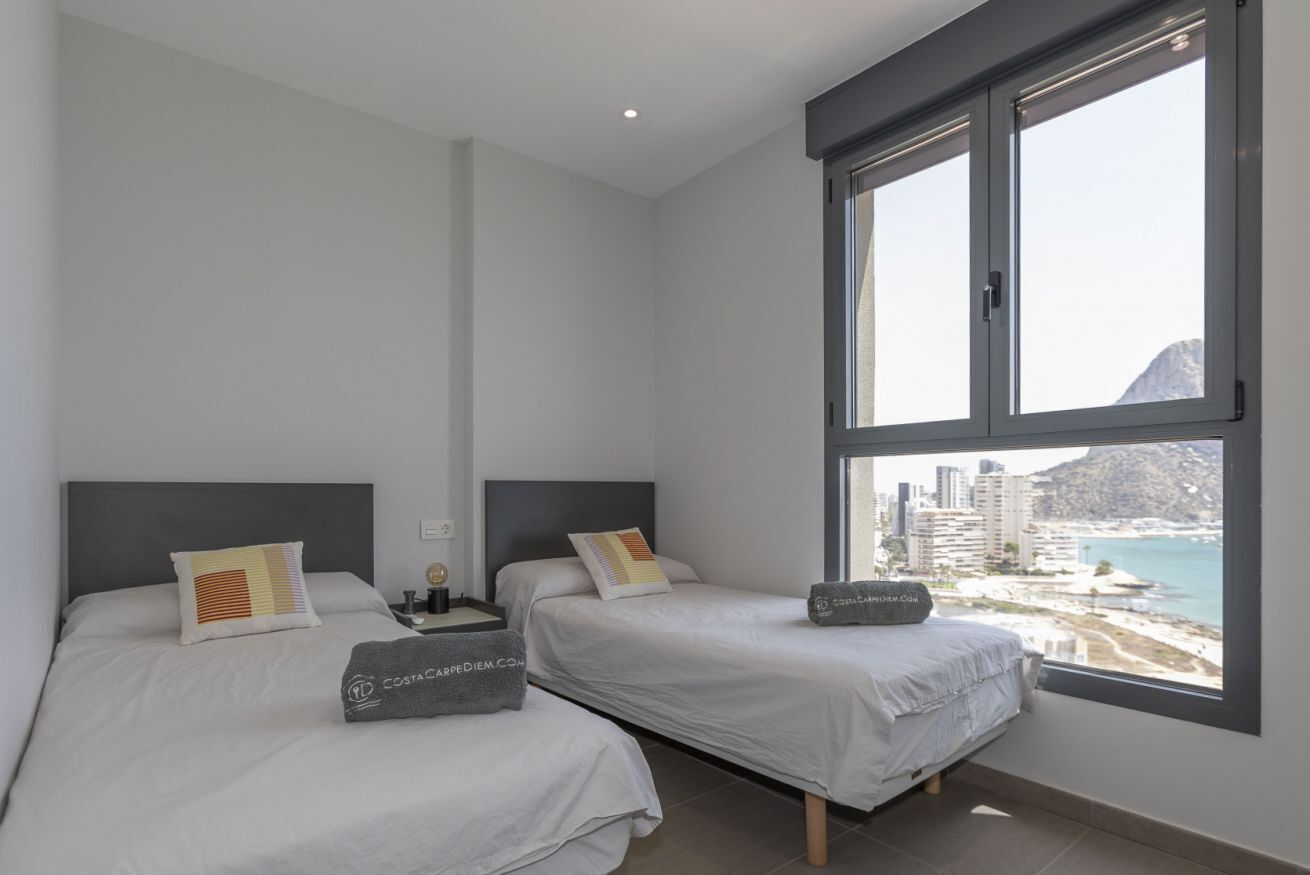 Moderni hotellihuone, jossa on kaksi sänkyä, suuri ikkuna, josta on näkymä kaupunkiin, sekä pieni pöytä, jossa on lamppu.