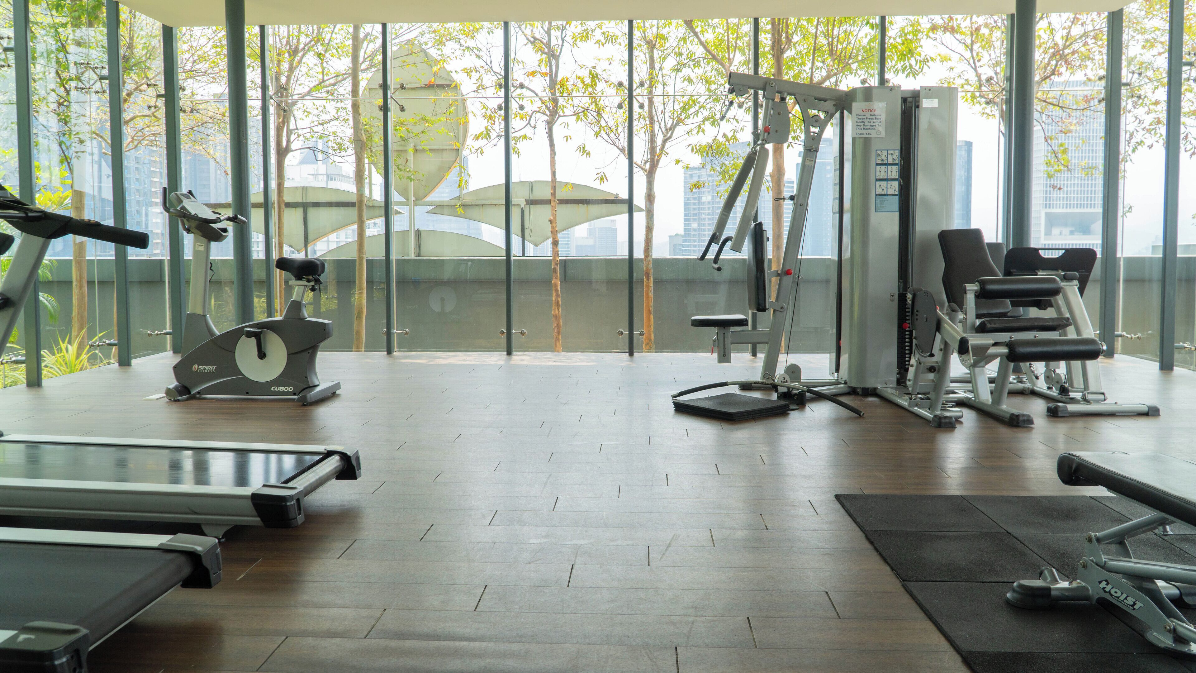 Un gimnasio con diferentes equipos de ejercicio, amplias ventanas y vistas a edificios y árboles.