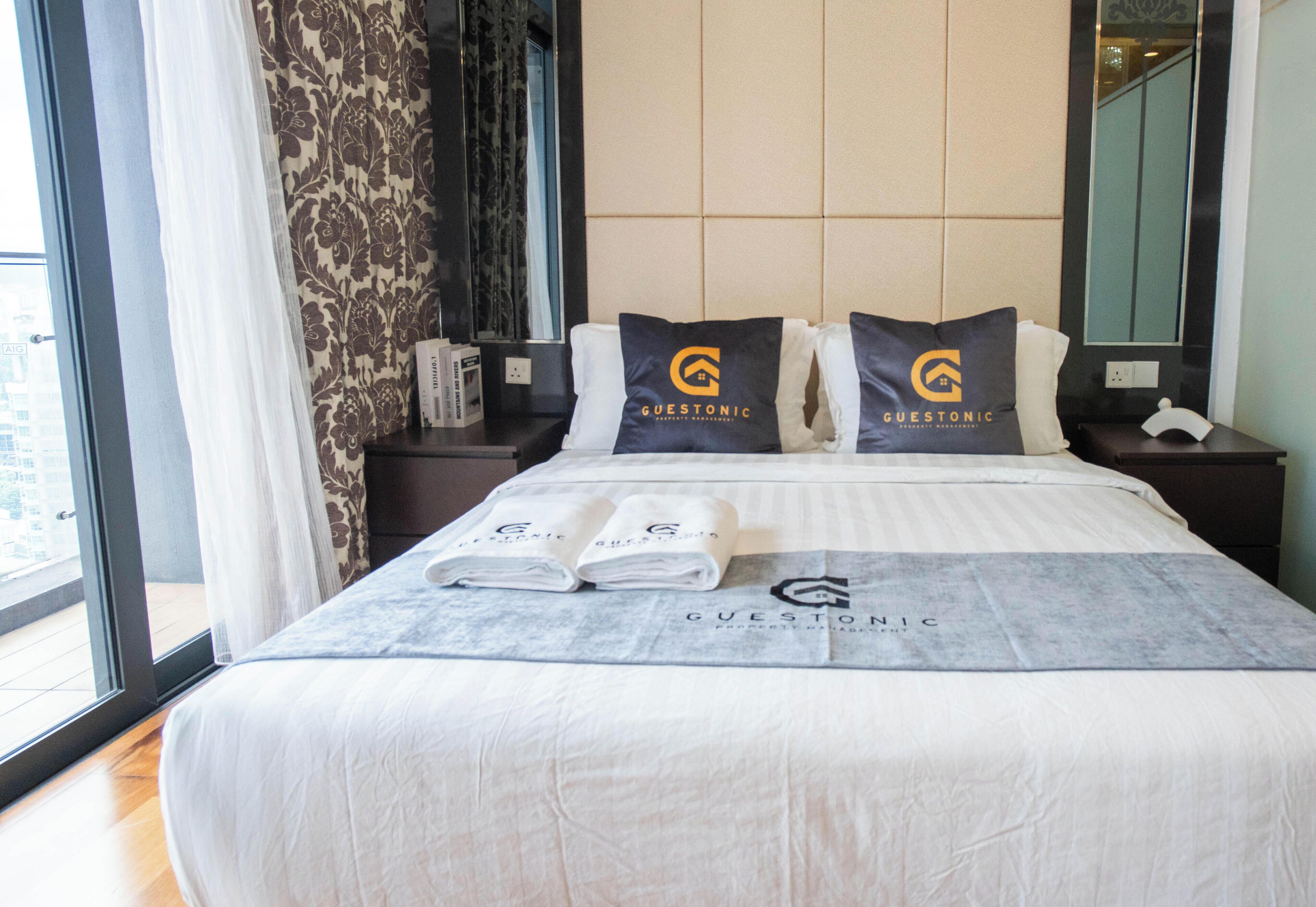 A hotel room with a large bed, two pillows with the logo 'Guestonic', a bedside table with a book and a phone, and a window with curtains.