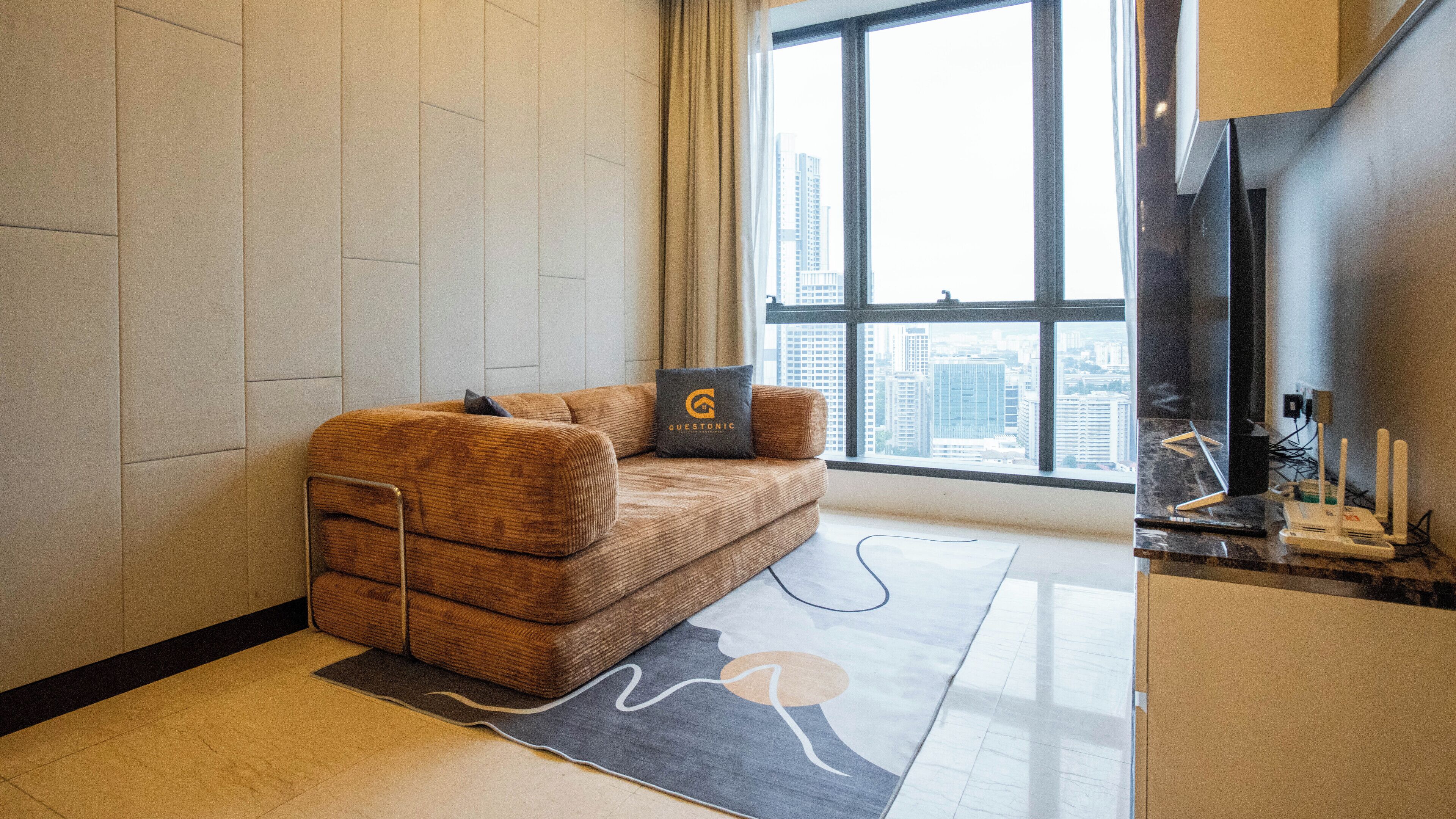 A modern hotel room with a brown sofa, a view of the city through large windows, and a small kitchenette area.