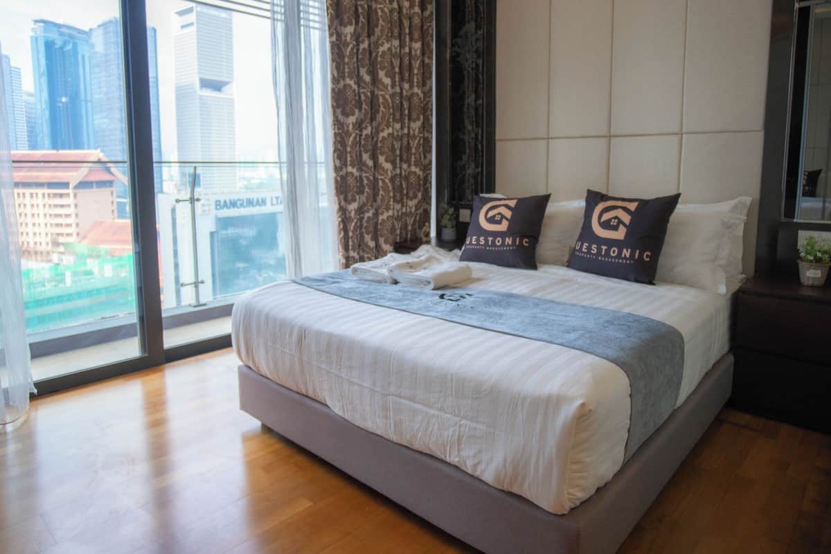 A hotel room with a large bed, two pillows with a logo, a nightstand with a plant, and a view of buildings through large windows.