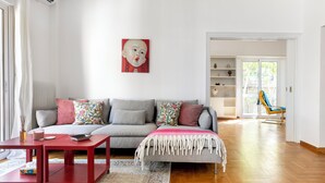A modern living room with a grey sofa, red coffee table, and a colorful throw blanket. There is a painting on the wall and a blue chair in the background. - Artsi - 3BR Apt w Balcony in Athens (Athens)