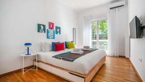 A modern bedroom with a wooden floor, a bed with colorful pillows, a small side table, and artwork on the wall. - Artsi - 3BR Apt w Balcony in Athens (Athens)