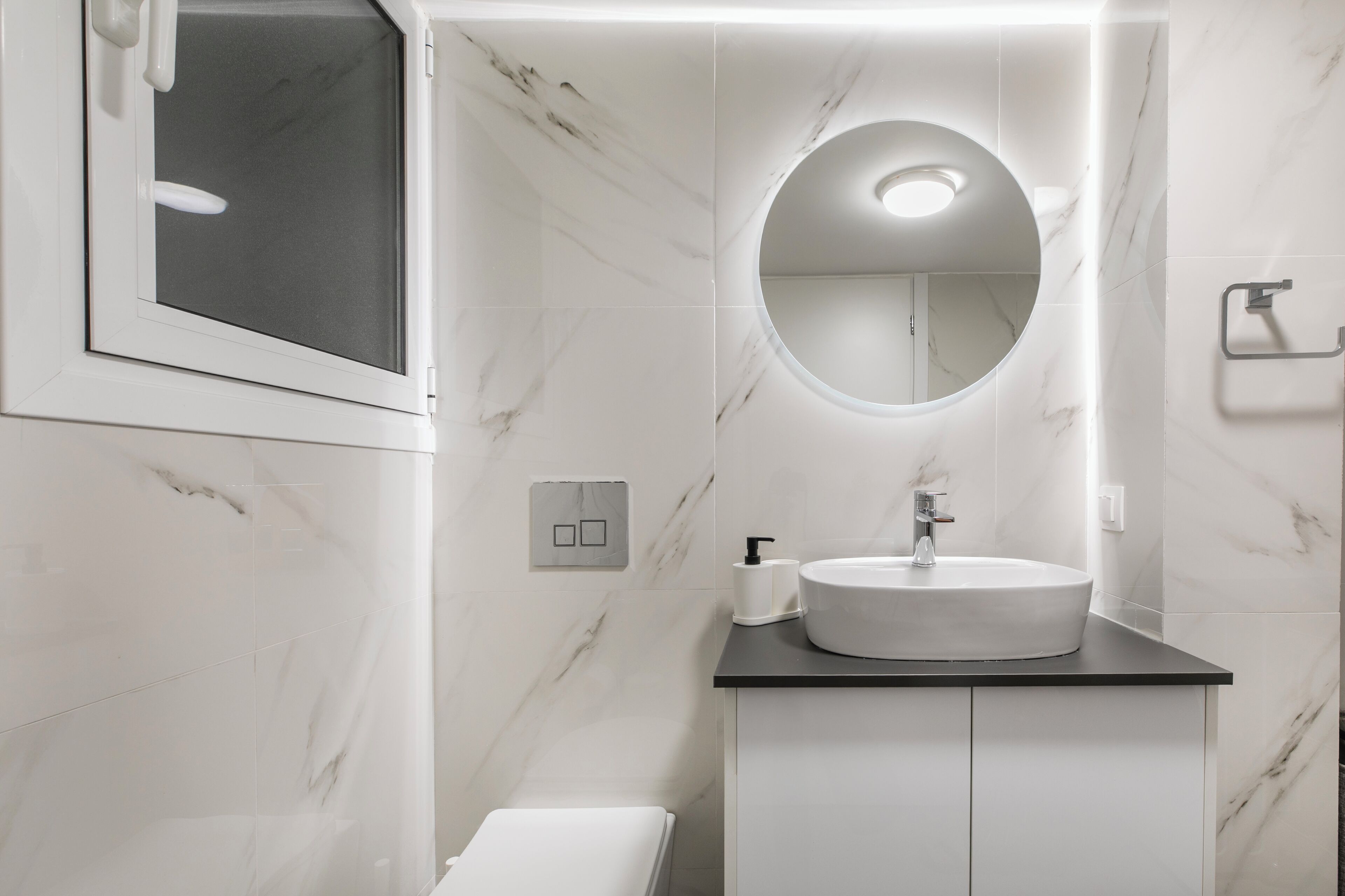 A modern bathroom with a white marble wall, a round mirror, a white sink, and a black countertop.