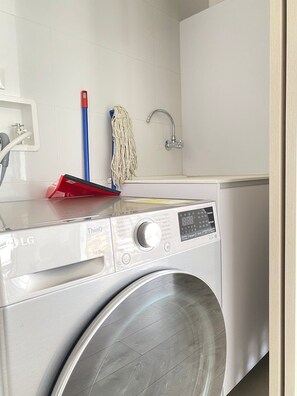A front view of a modern laundry room with a front-loading washing machine, a mop, a broom, and a sink. - Edificio Area 93  (Bogotá)