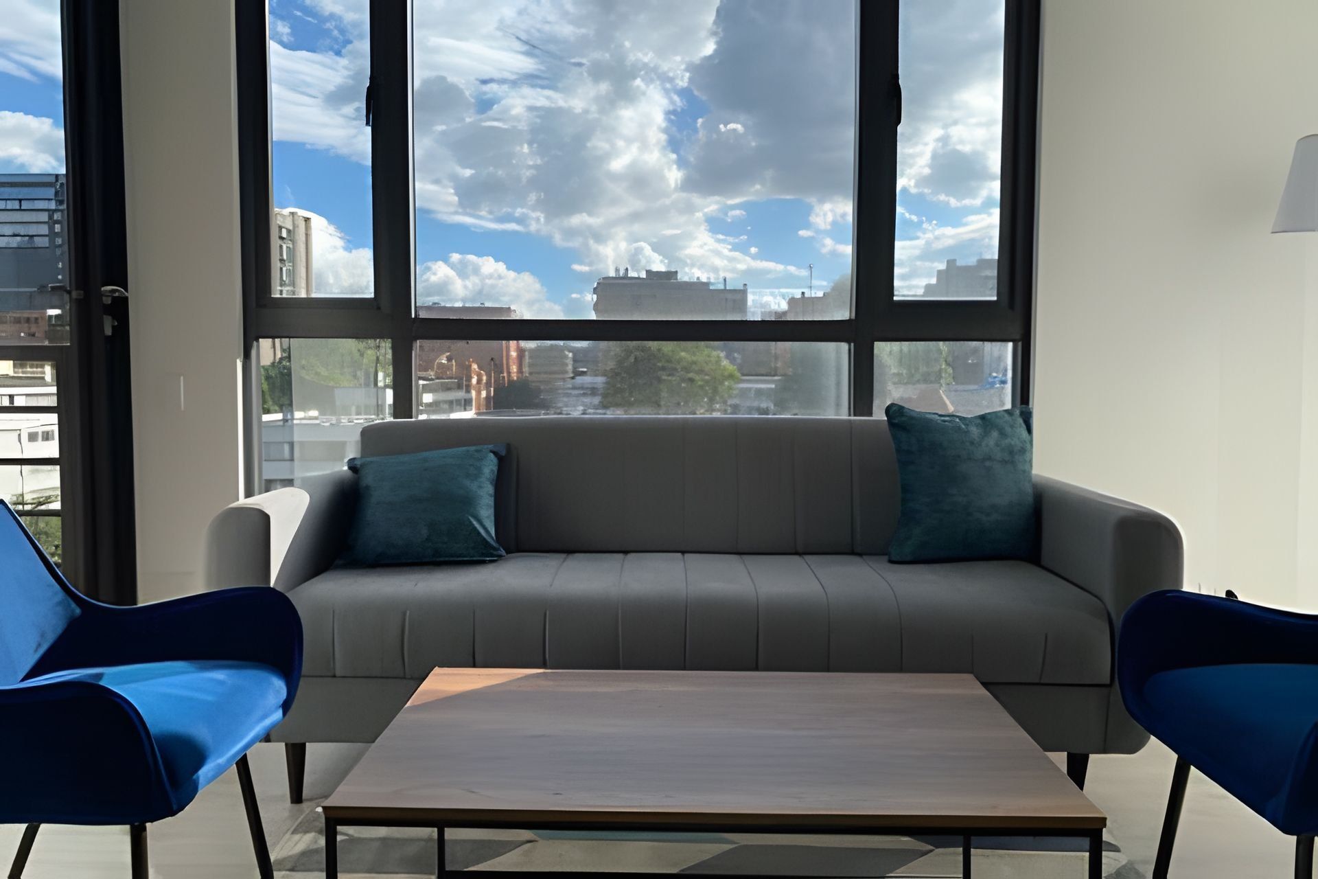 Une pièce moderne avec un canapé gris, deux fauteuils bleus et une table en bois. De grandes fenêtres offrent une vue sur le paysage urbain.