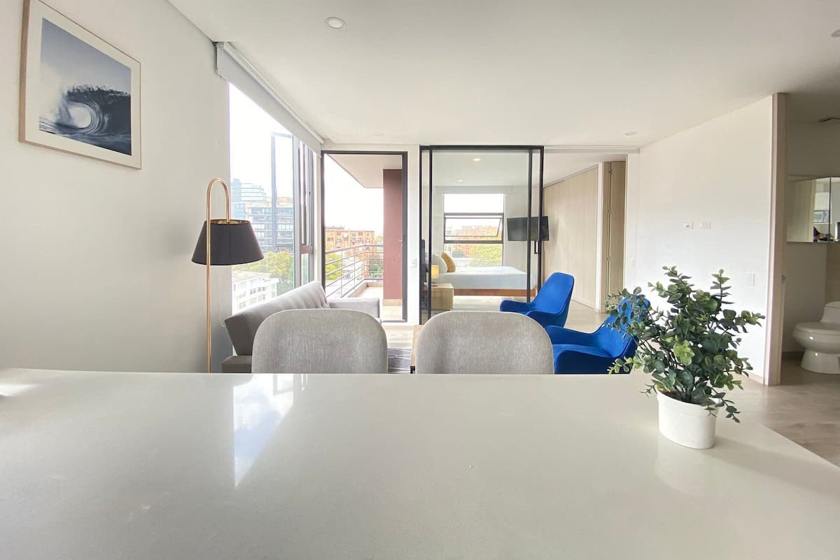 Una sala de estar moderna con una mesa grande, dos sillas, una planta en maceta y vistas de la ciudad a través de amplios ventanales.
