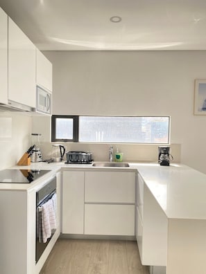 A modern kitchen with white cabinetry, a built-in oven, a microwave, a coffee maker, and a window providing natural light. - Edificio Area 93  (Bogotá)