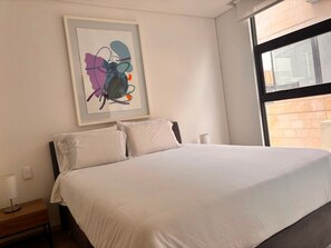 A hotel room with a bed, a nightstand, a lamp, and a window with a view of a brick wall. - Edificio Area 93  (Bogotá)