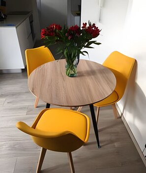 A dining area with a round wooden table, two yellow chairs, and a vase with red flowers.