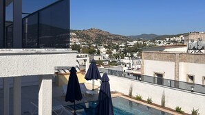 Ein Pool auf dem Dach mit Liegestühlen und Sonnenschirmen, mit Blick auf eine Stadt mit weißen Gebäuden und einer hügeligen Landschaft.