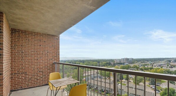 Ein Balkon mit Backsteinmauer, zwei gelbe Stühle, einem Holztisch und Blick auf einen Parkplatz und Gebäude.