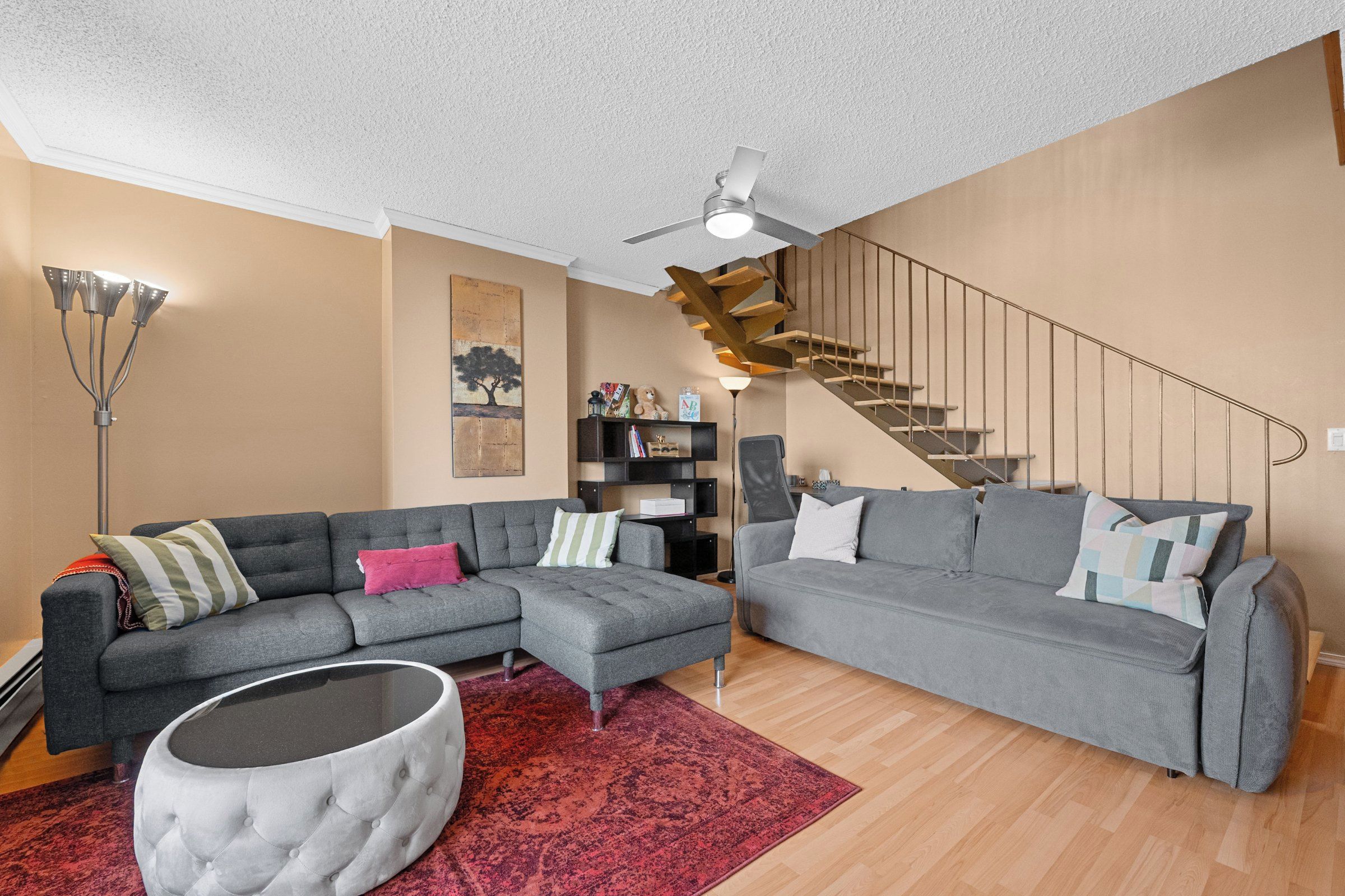 A modern living room with two grey sofas, a red patterned rug, a round ottoman, a ceiling fan, and a staircase.