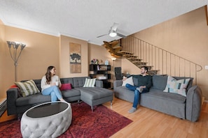 Uma sala de estar moderna com dois sofás cinzentos, um tapete estampado em vermelho e uma escada com guarda-corpos de madeira.