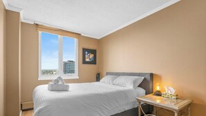 A bedroom with a bed, a bedside table with a lamp, a window with blinds, and a framed picture on the wall.