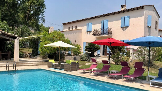 A swimming pool with lounge chairs and umbrellas, a stone wall, and a building with blue shutters. - BASTIDE DE L'ORANGERAIE SWIMMING POOL AIR CONDITIONING (Vence)
