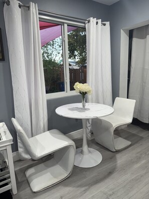 A room with a white table and two chairs, a window with curtains, and a vase with flowers on the table.