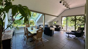Een ruime kamer met een eettafel, stoelen en een bank. Een groot raam zorgt voor daglicht en uitzicht op de omgeving.