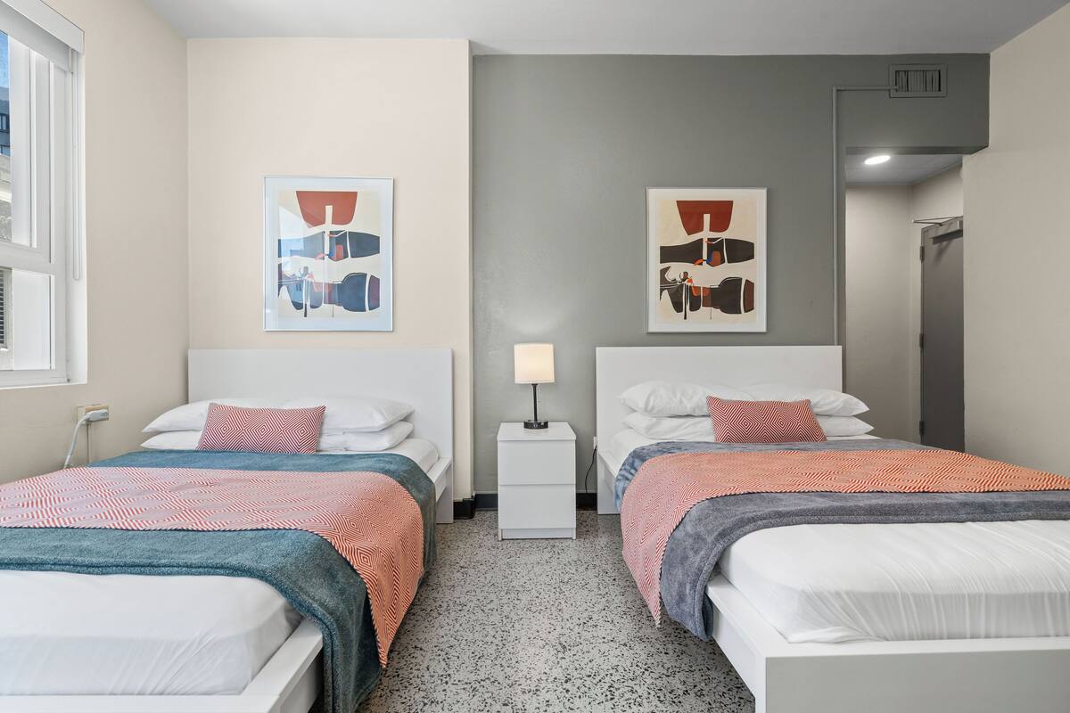 A hotel room with two beds, a nightstand, a lamp, and abstract artwork on the walls.