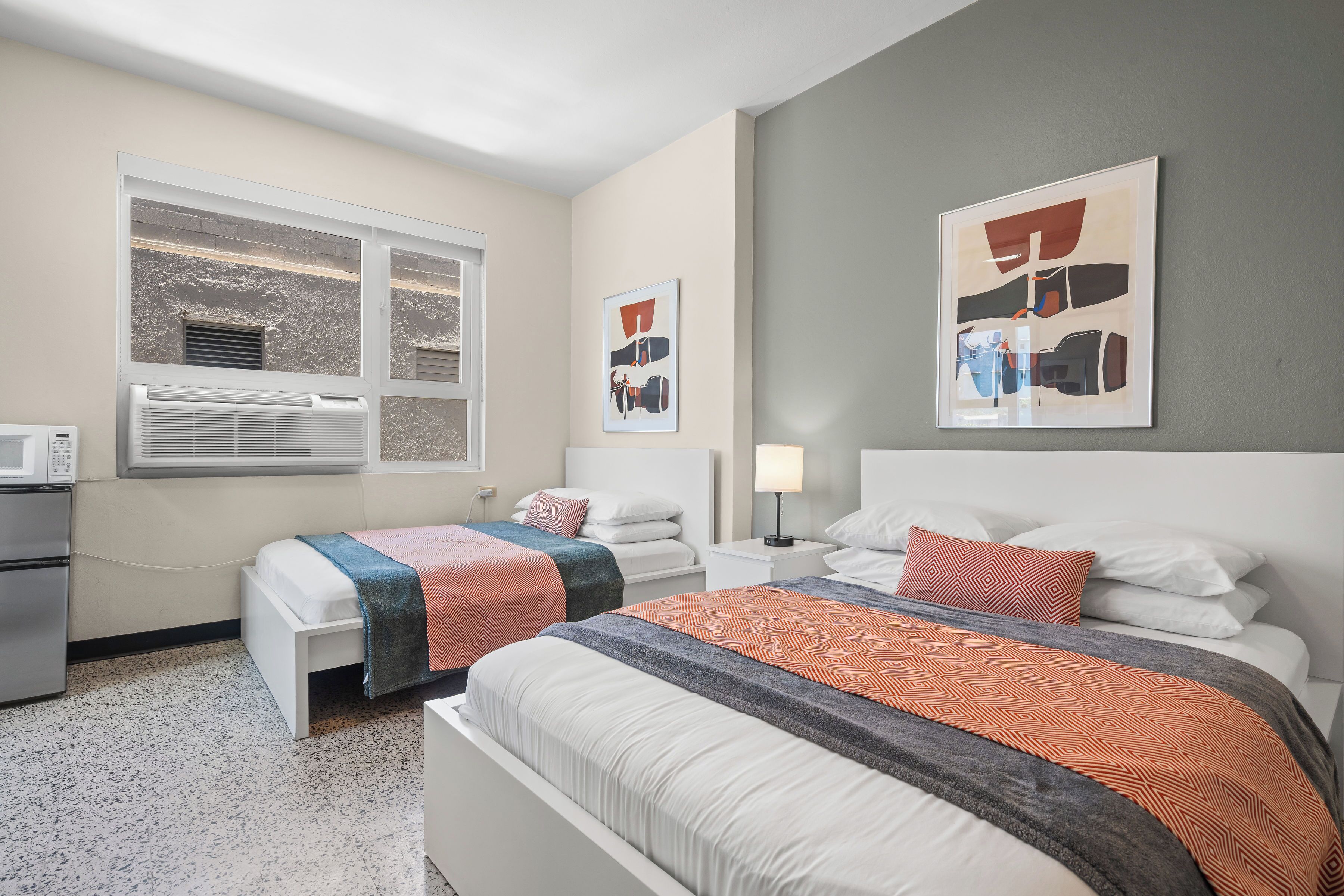 A hotel room with two beds, a kitchenette, a window with blinds, and artwork on the walls.