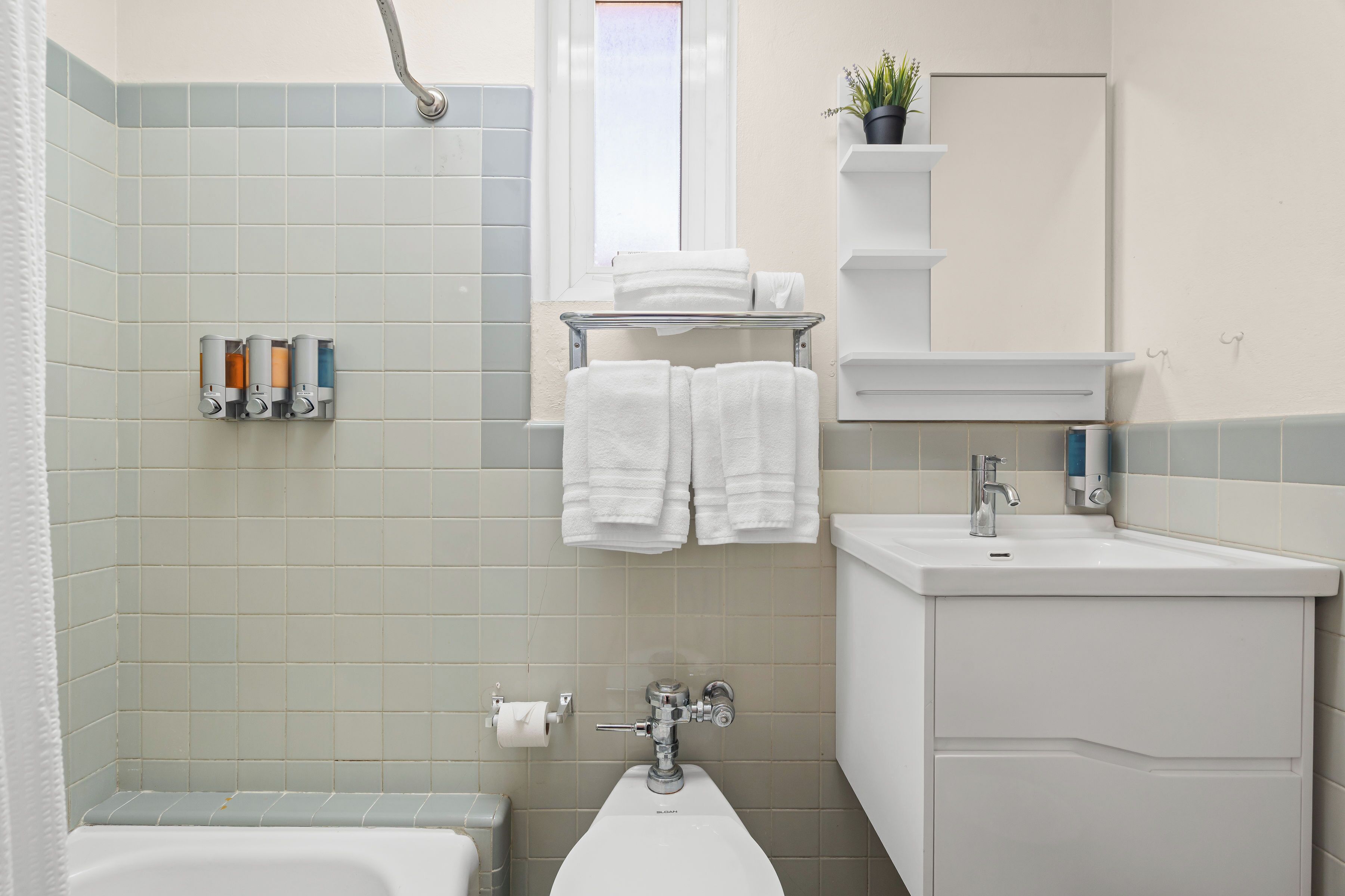 A bathroom with a bathtub, a sink, a toilet, a towel rack with towels, a shower curtain, a window, and a potted plant on a shelf.