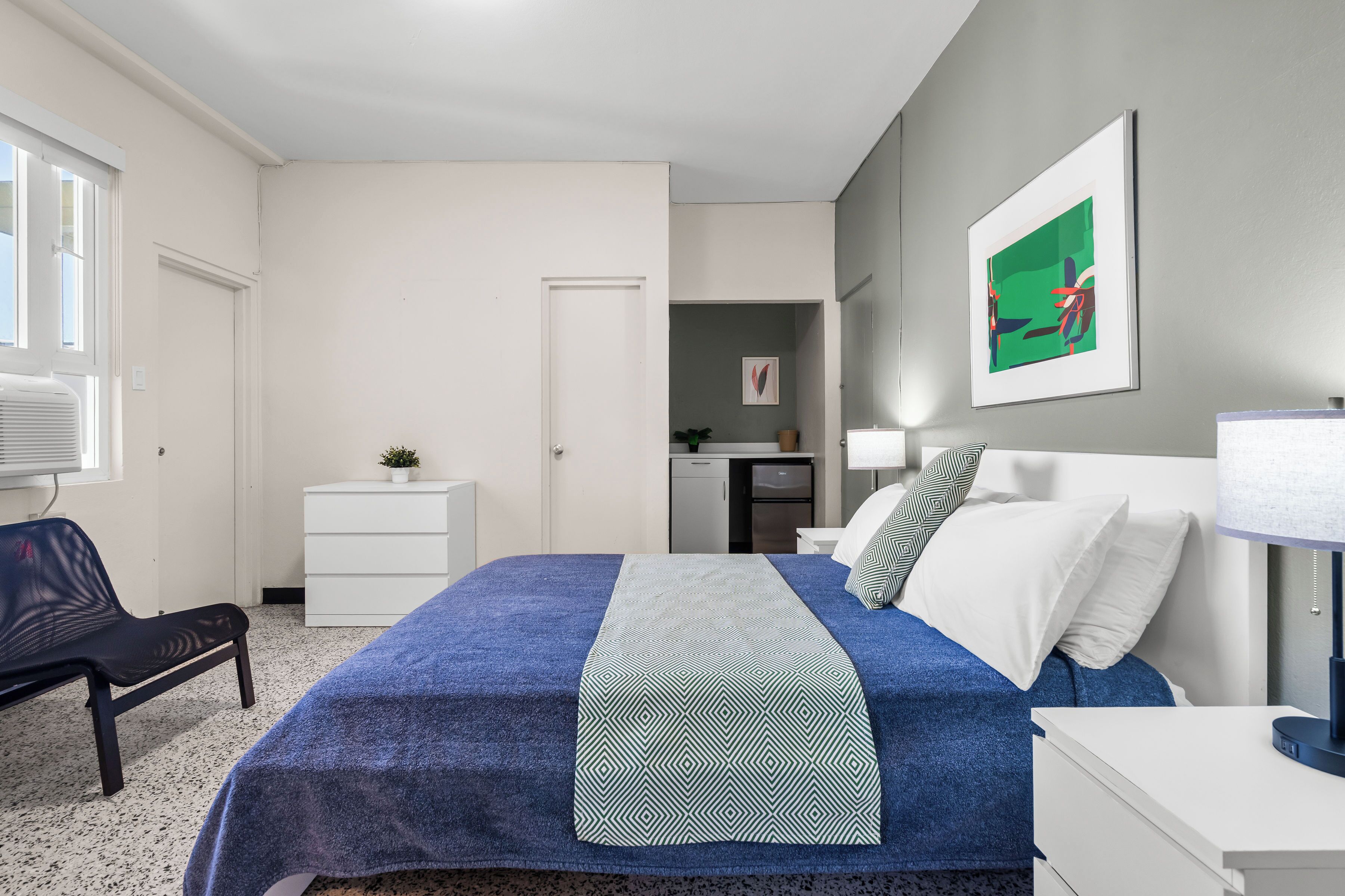 A bedroom with a bed, a bedside table, a chair, a desk, and a small kitchenette in the background.