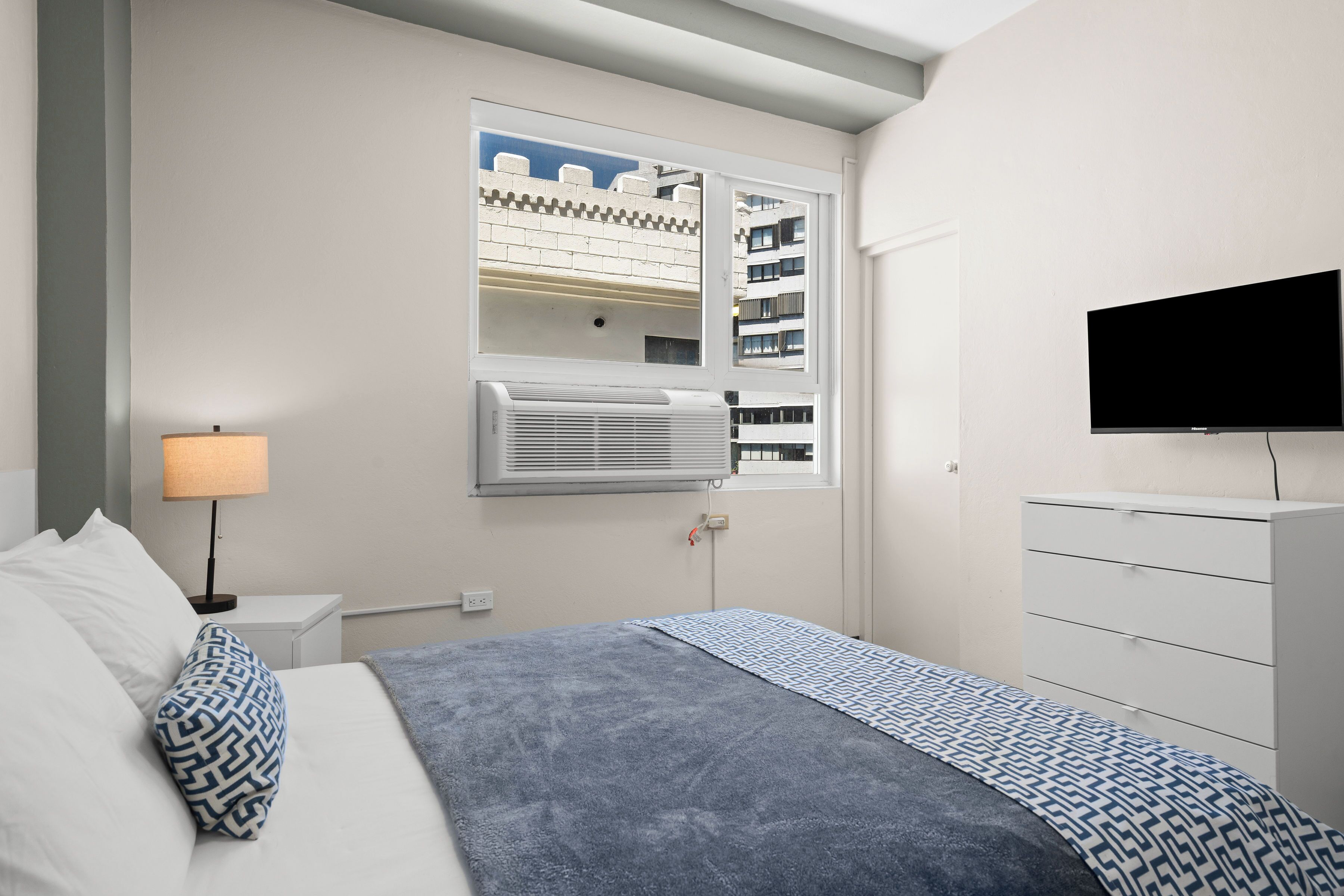 Ein Schlafzimmer mit einem Bett, einem an der Wand befestigten Fernseher, einem Fenster mit Blick auf Gebäude und einem Nachttisch mit Lampe.