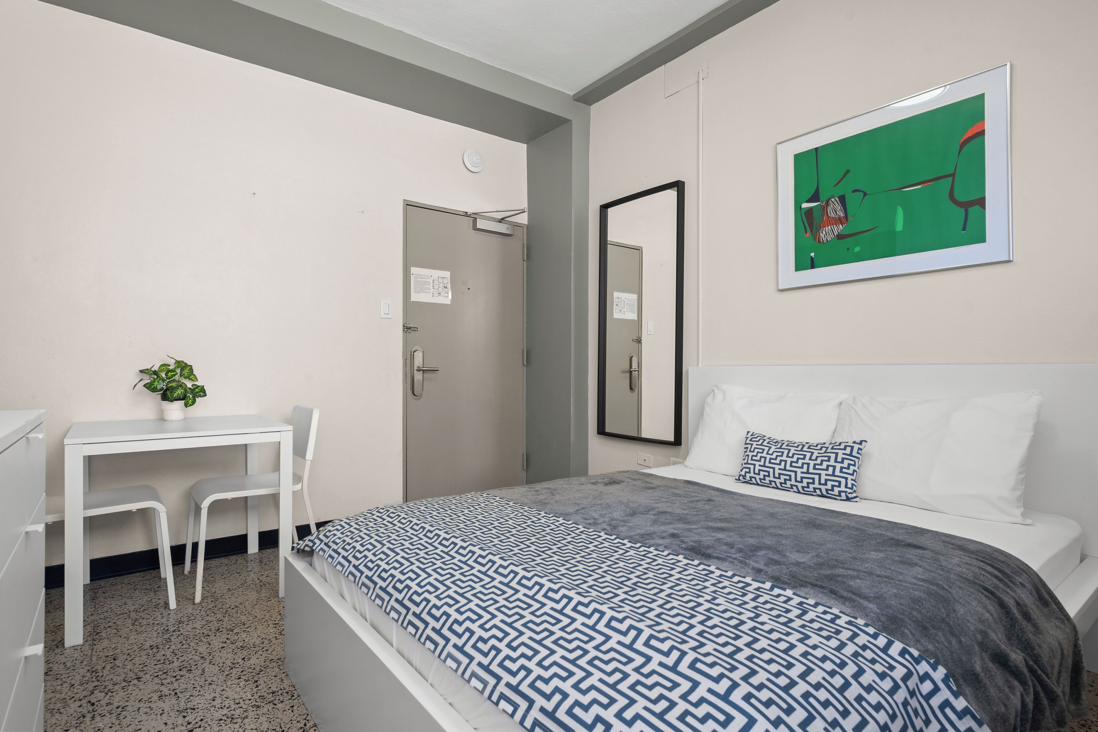 Une chambre à coucher avec un lit, un bureau et une chaise. Il y a un miroir au mur et un tableau au-dessus du lit.