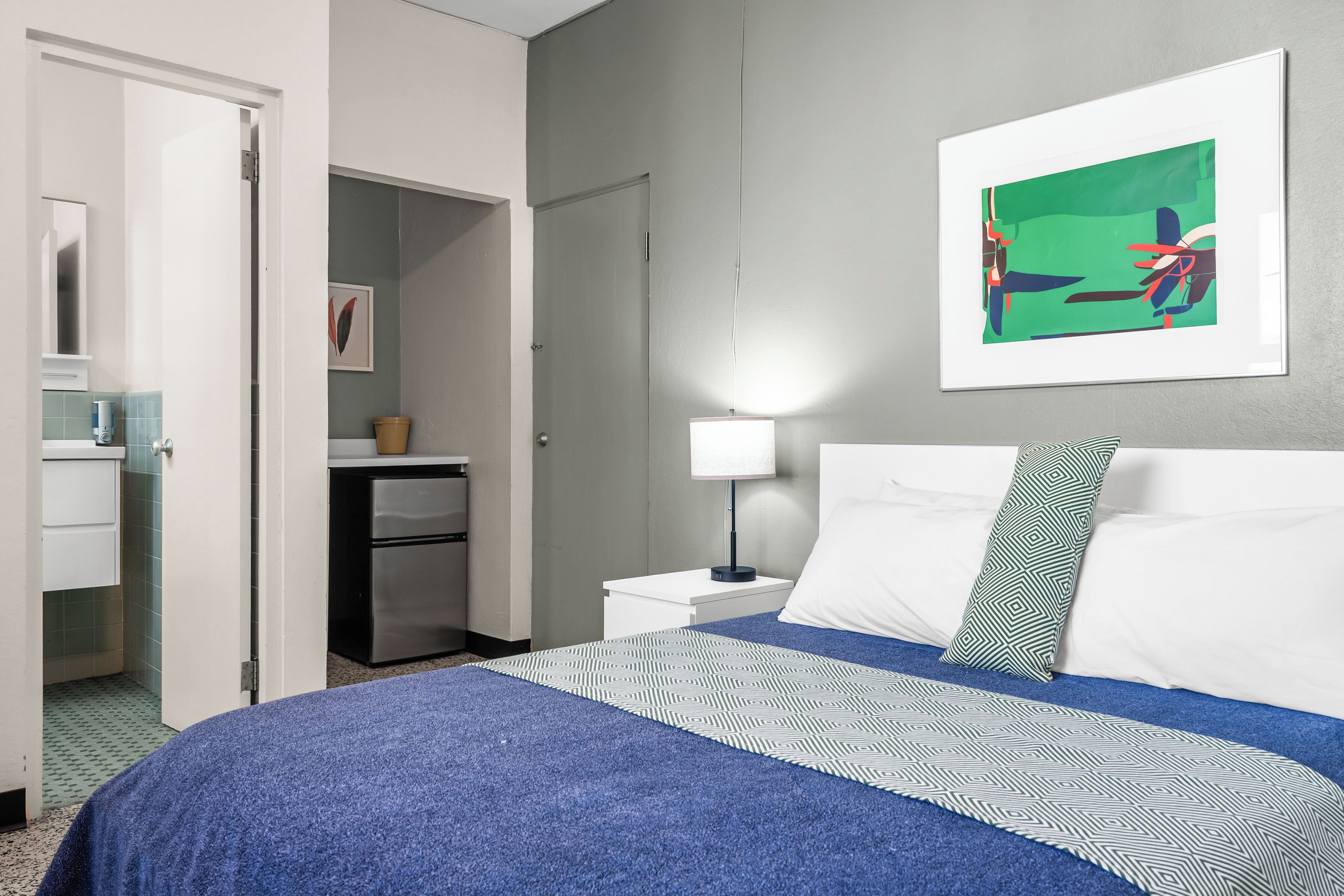 Habitación de hotel con cama, mesita de noche, lámpara, un cuadro en la pared y un baño visible a través de una puerta abierta.