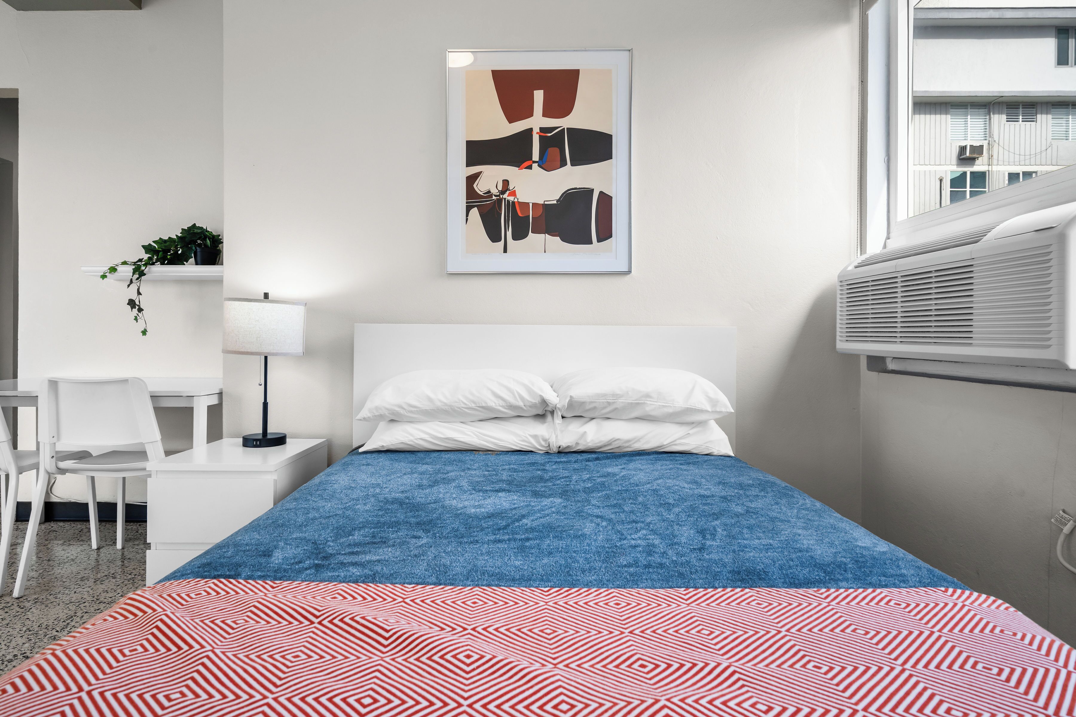 A neatly arranged hotel room with a bed, a desk, a chair, a lamp, and a wall-mounted air conditioner.