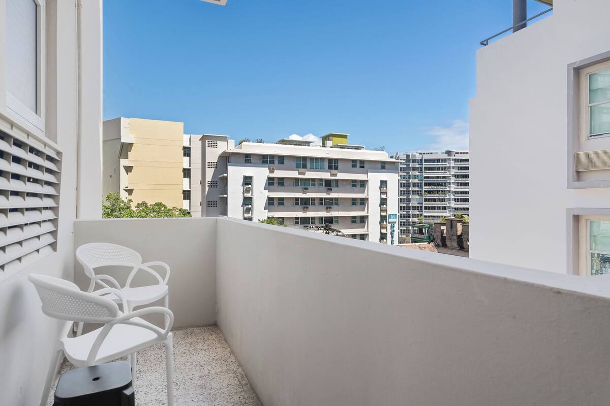 A balcony with white chairs and a small table, overlooking a cityscape with modern buildings.