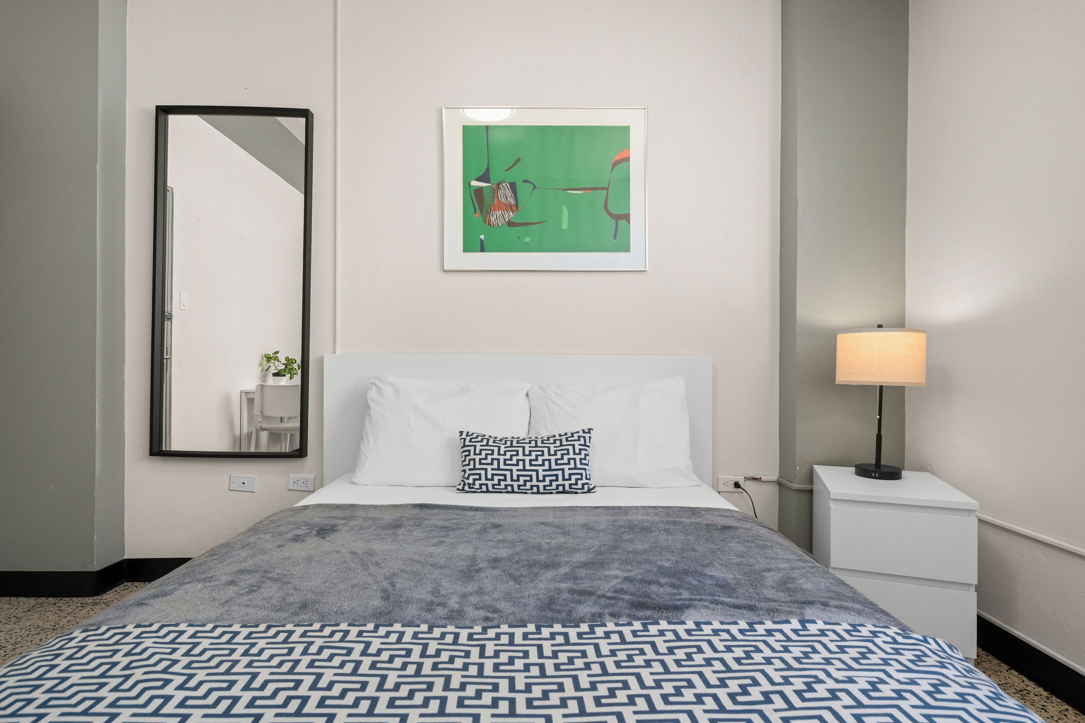 A hotel room with a bed, a nightstand with a lamp, a framed picture on the wall, and a mirror.