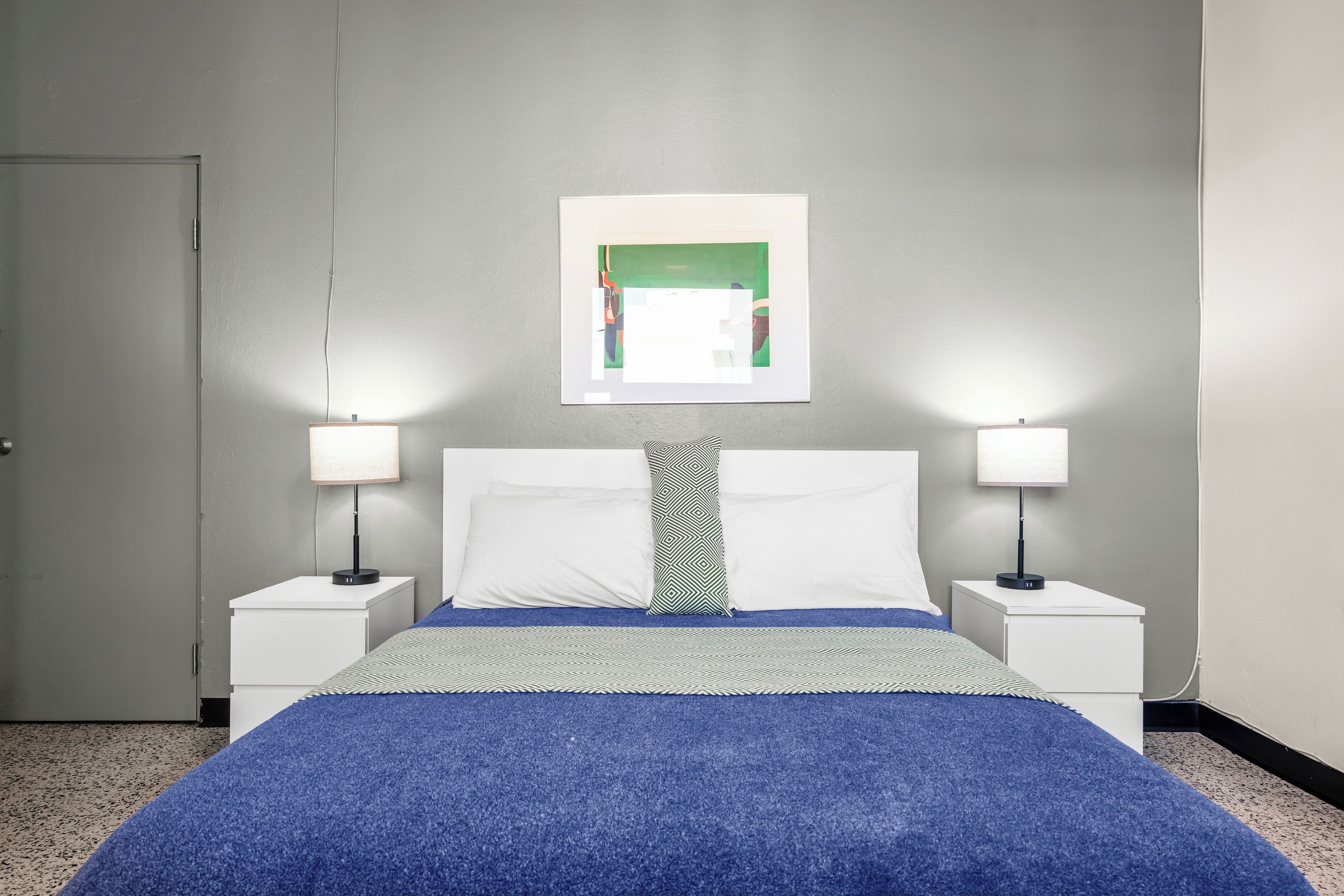 A neatly arranged hotel room with a bed, two bedside tables with lamps, a door, and a framed picture on the wall.