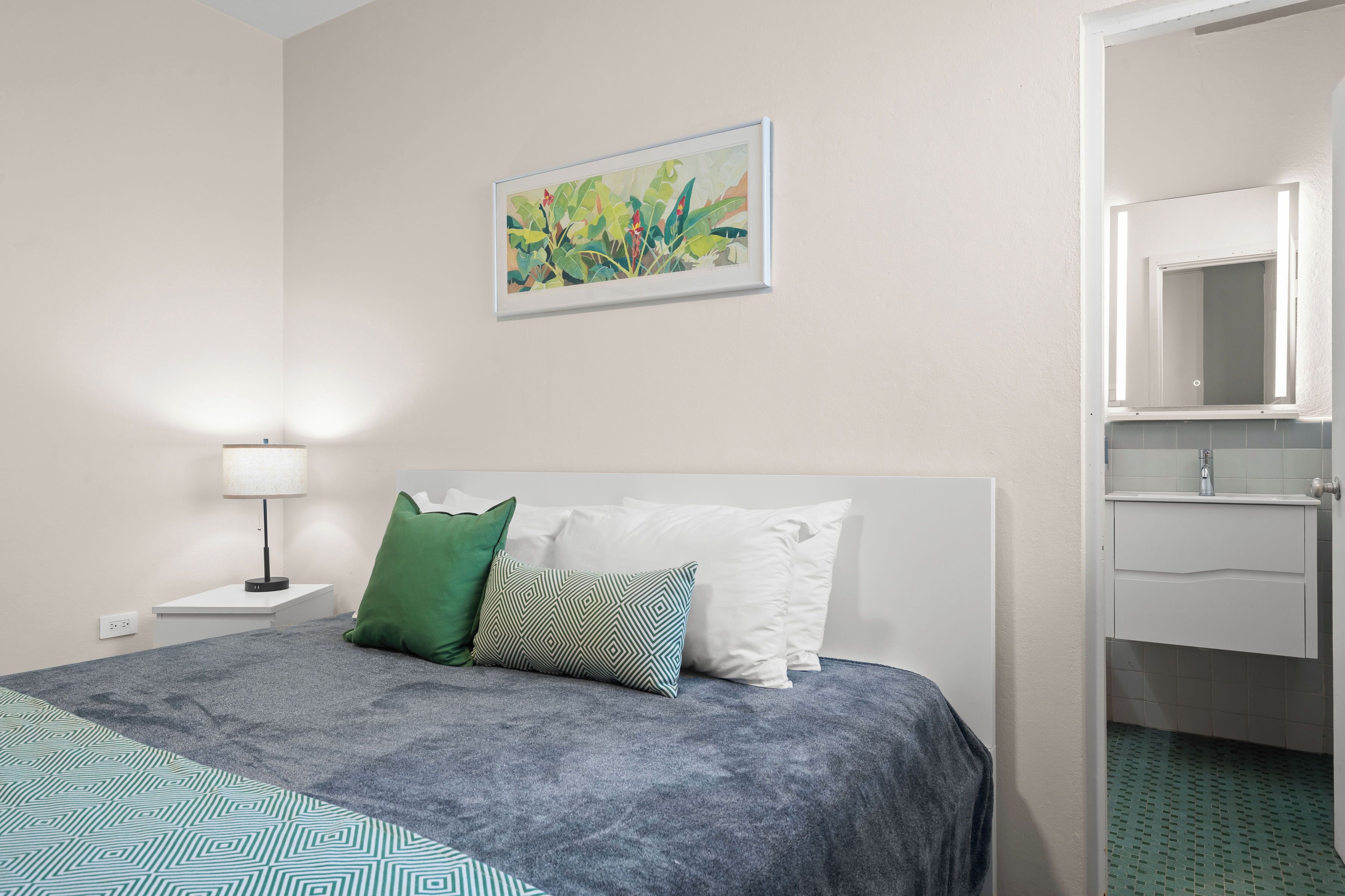Habitación de hotel ordenada con cama, mesita de noche, lámpara, un cuadro y un baño visible a través de una puerta abierta.