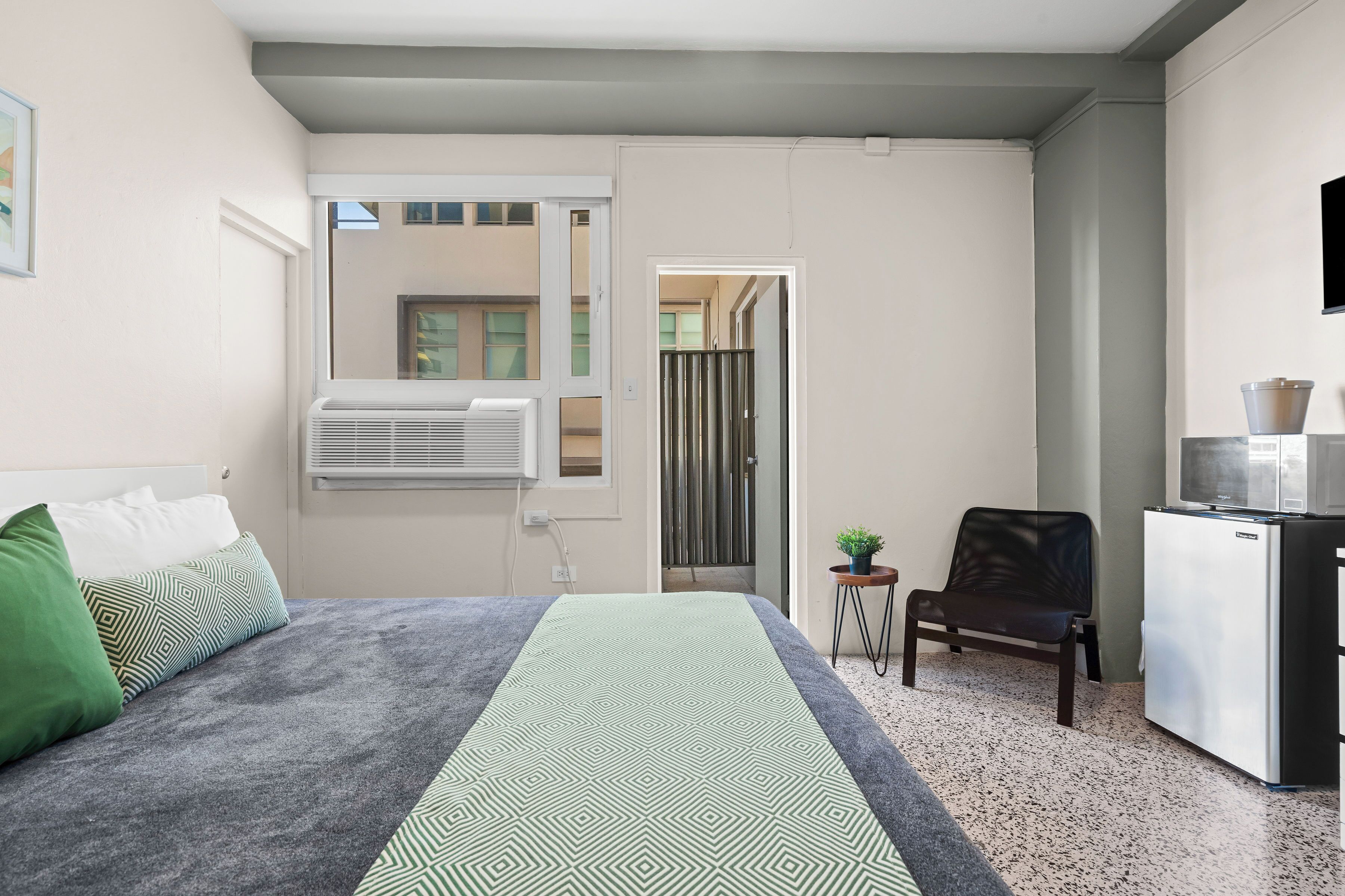 Un dormitorio con cama, aire acondicionado, una mesita con una planta, una silla y un refrigerador.