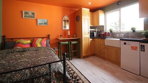 Une chambre à coucher de taille modeste, comprenant un lit, un petit bureau et une fenêtre. La pièce adjacente est une cuisine équipée de meubles en bois, d’un évier et d’un réfrigérateur.