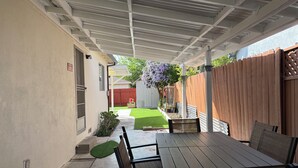 Ein überdachter Patio-Bereich mit Tisch und Stühlen, ein kleiner Garten mit Rasen und einem Holzzaun sowie ein Schuppen im Hintergrund.