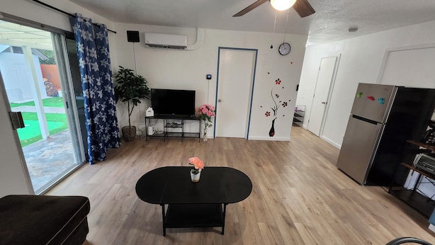 Ein modernes Wohnzimmer mit Flachbildfernseher, einem Esstisch mit Blumenvase, Deckenventilator und Kühlschrank.