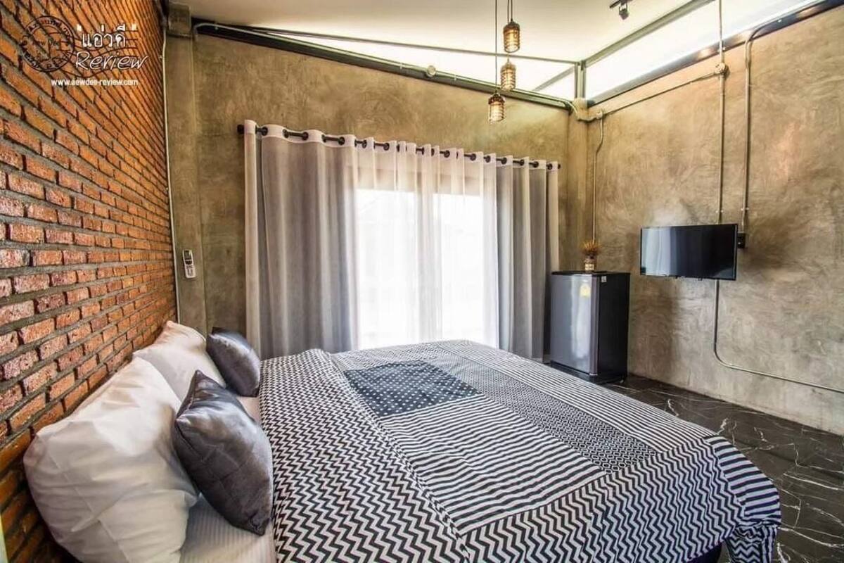 Un dormitorio con pared de ladrillo, una cama con ropa de cama estampada, un televisor y una ventana con cortinas.